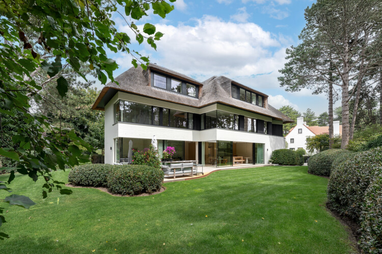 Luxueuze villa te koop in Knokke met witte gevel, rieten dak en aangelegde tuin met gazon.